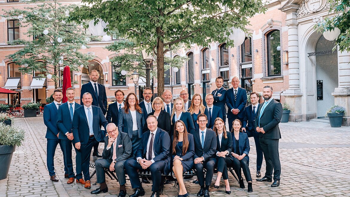 Das Team von Solvium Capital GmbH posiert in Anzügen im Freien auf einem gepflasterten Platz vor historischen Gebäuden; einige sitzen auf einer Bank, andere stehen dahinter, alle lächeln in die Kamera.