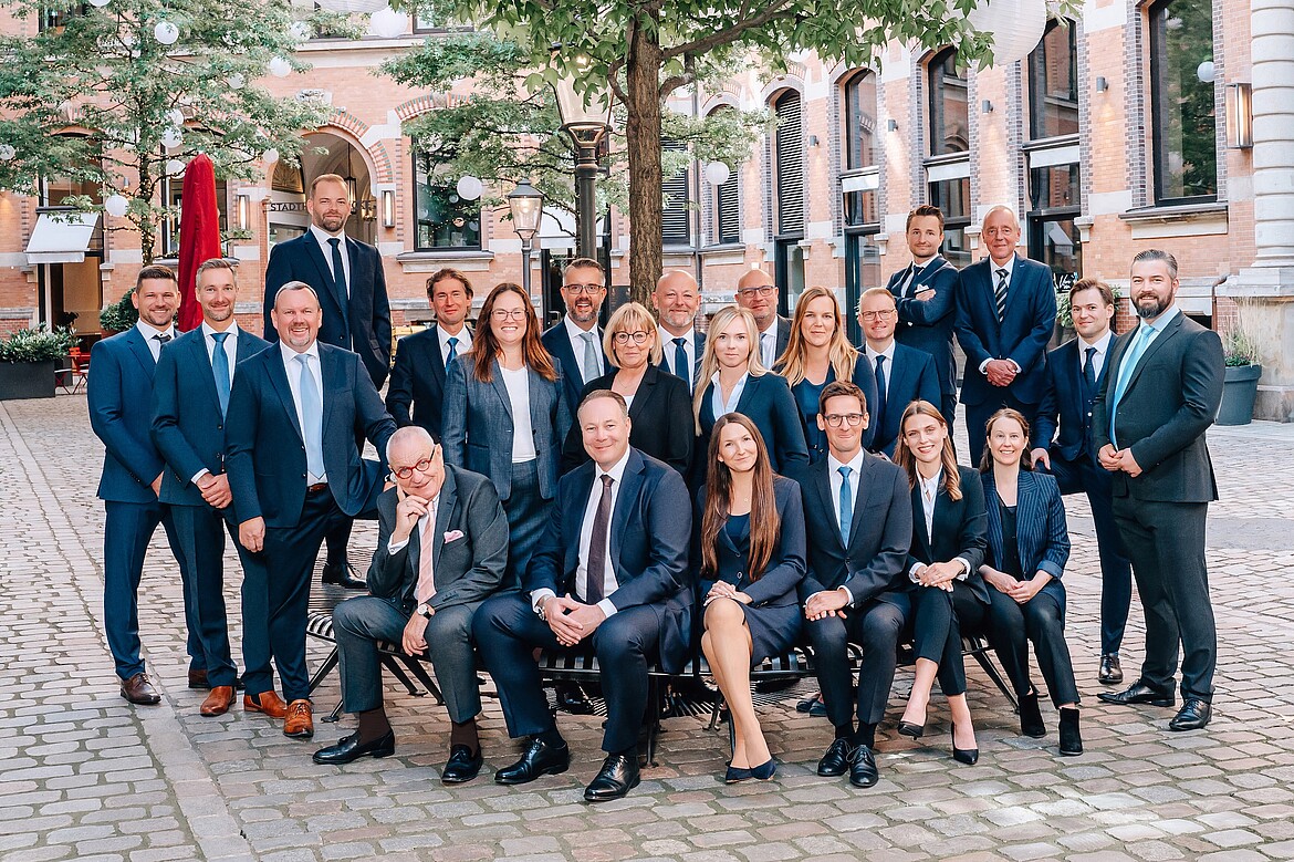 Das Team von Solvium Capital GmbH posiert in Anzügen im Freien auf einem gepflasterten Platz vor historischen Gebäuden; einige sitzen auf einer Bank, andere stehen dahinter, alle lächeln in die Kamera.